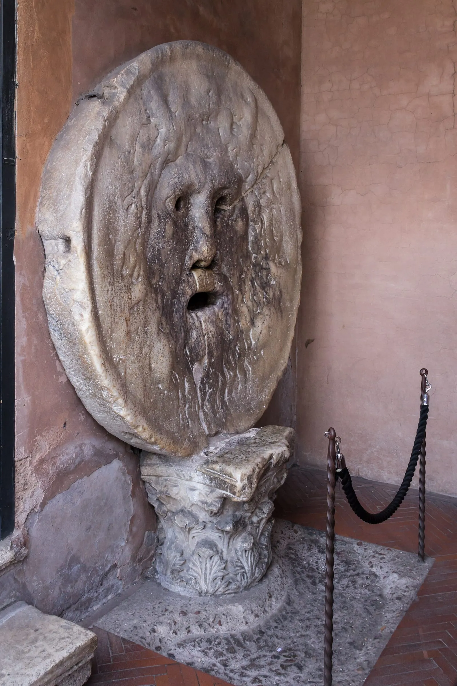 Le disque de marbre de la Bocca della Verità, visage barbu à la bouche ouverte, adossé au mur du portique de Santa Maria in Cosmedin et posé sur un chapiteau corinthien