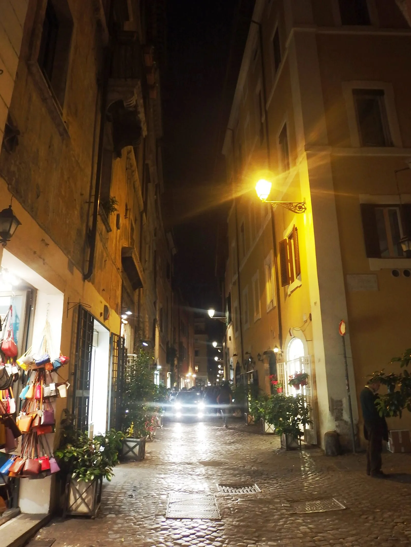 Ruelle du centre historique de Rome la nuit, chaussée pavée de sampietrini, façades ocre, lampadaire mural en fer forgé et vitrine de maroquinerie éclairée