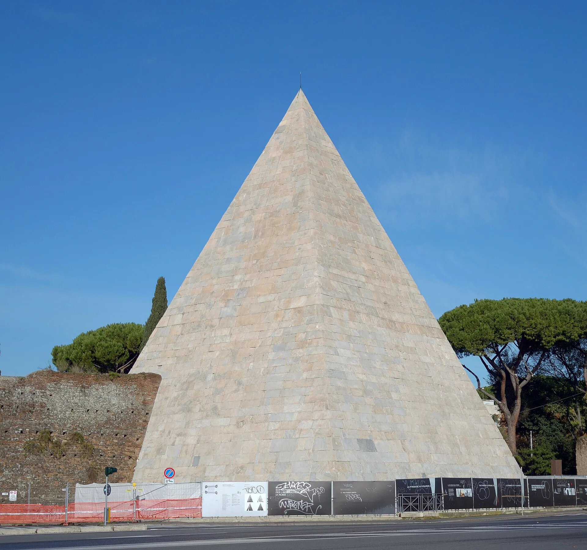 La pyramide de Cestius à Rome, en pierre claire, vue depuis la rue, avec le mur d'Aurélien et des pins à sa gauche