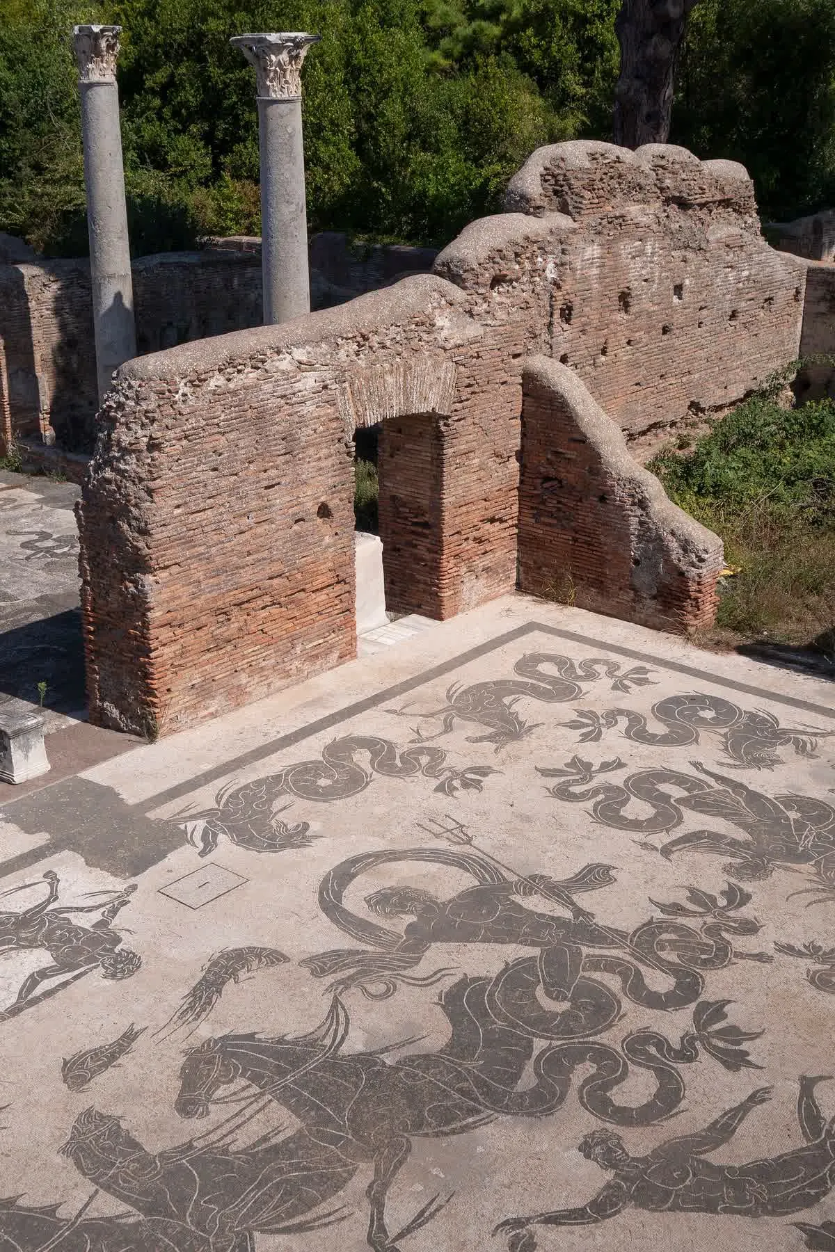 Mosaïque en noir et blanc des thermes de Neptune à Ostie Antica, avec figures marines et chevaux, devant des murs de brique et deux colonnes