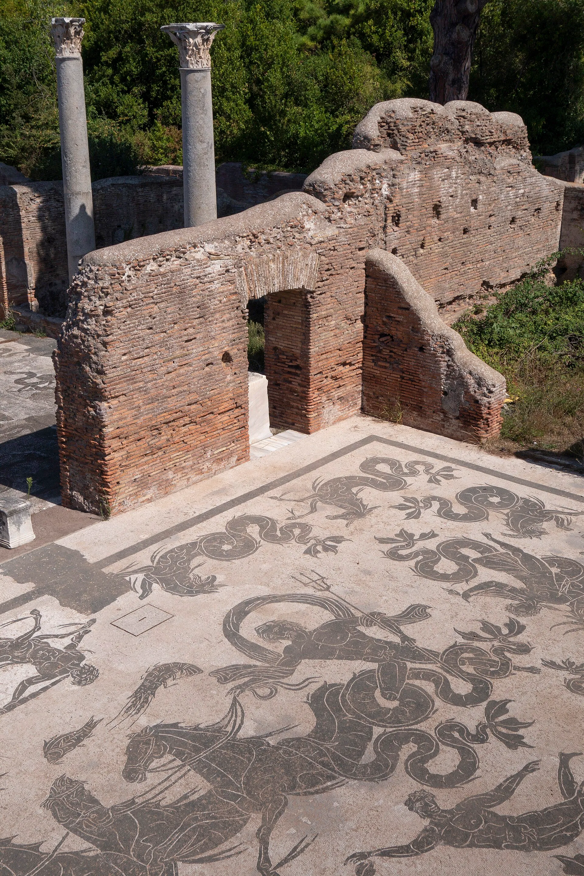 Mosaïque en noir et blanc des thermes de Neptune à Ostie Antica, avec figures marines et chevaux, devant des murs de brique et deux colonnes