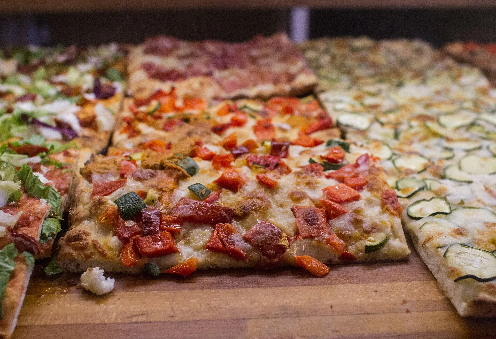 Parts de pizza al taglio aux légumes grillés et à la charcuterie, alignées sur une planche de bois