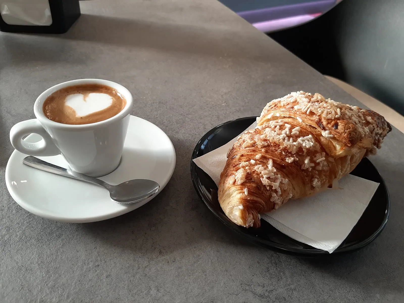 Un cappuccino dans sa tasse blanche posée sur une soucoupe, à côté d’un cornetto doré saupoudré de sucre et servi sur une petite assiette noire, sur le marbre d’un bar