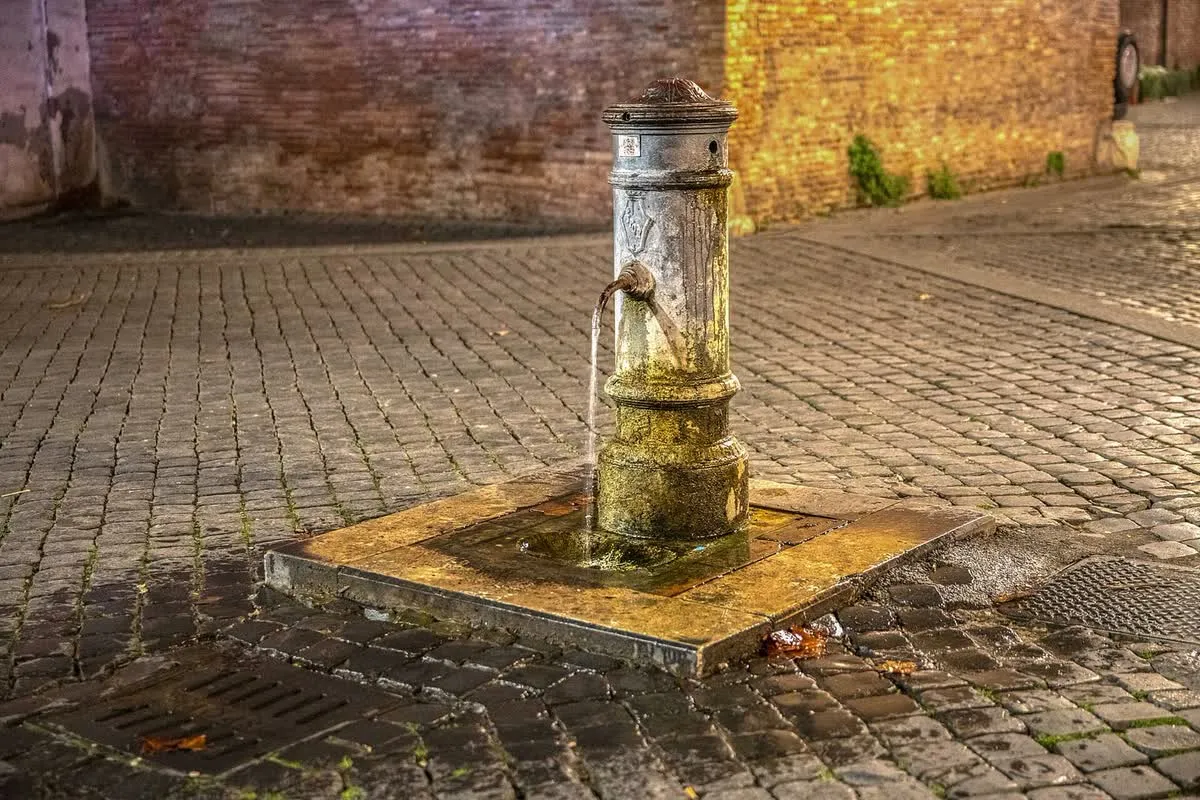 Un nasone en fonte au milieu d’une ruelle pavée de Rome, l’eau s’écoulant de son bec dans la lumière dorée du soir