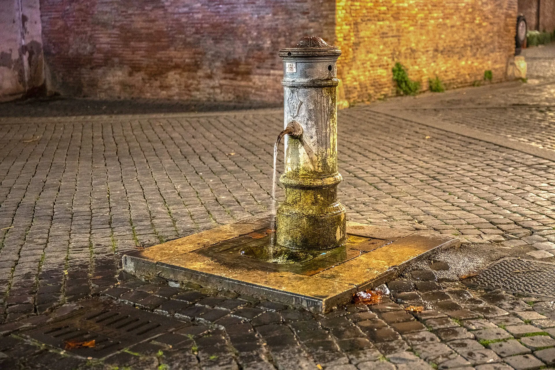 Un nasone en fonte au milieu d’une ruelle pavée de Rome, l’eau s’écoulant de son bec dans la lumière dorée du soir