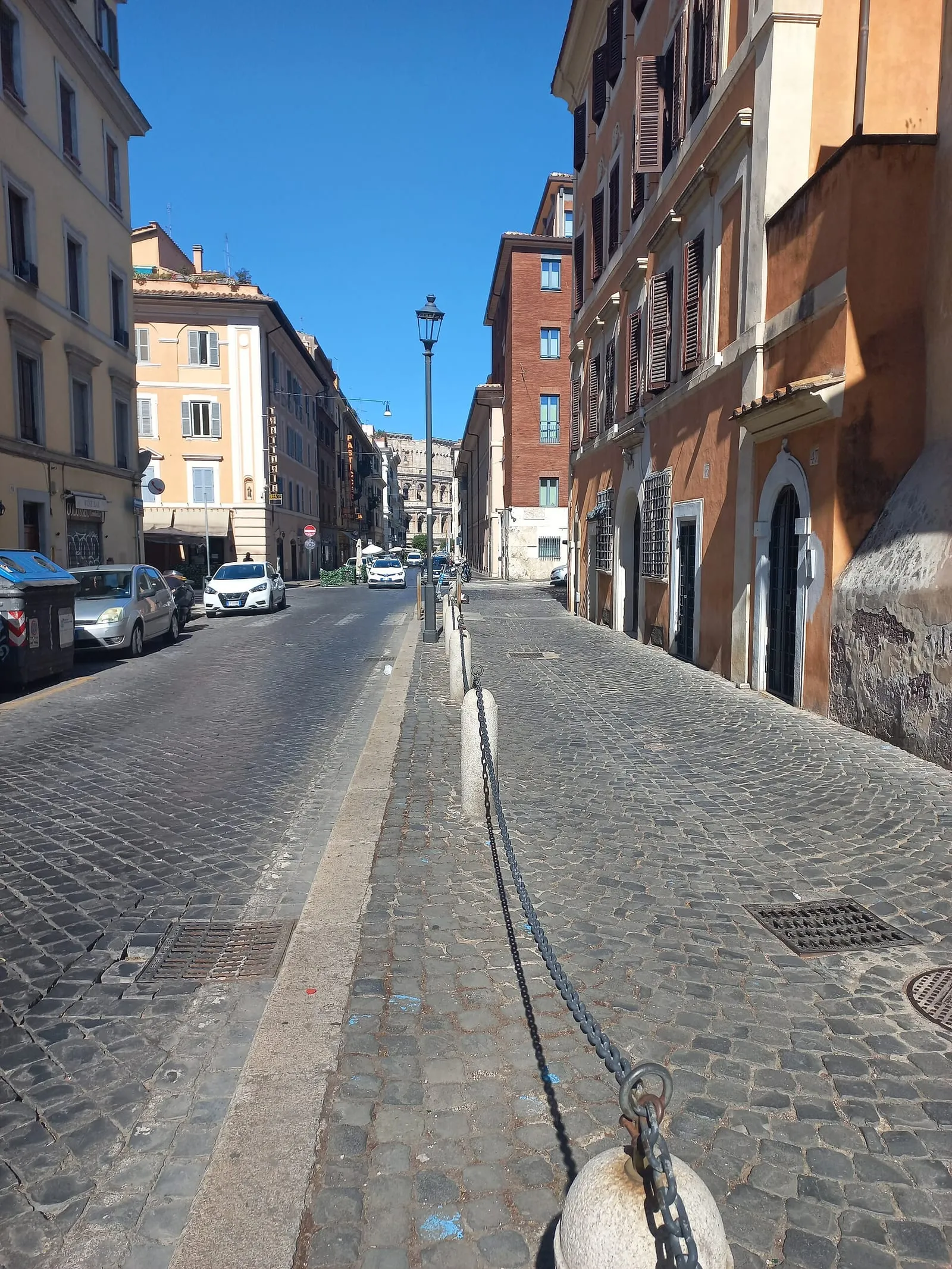 Rue du centre de Rome pavée en sampietrini, bordée de bornes et de chaînes, avec le Colisée au bout de la perspective