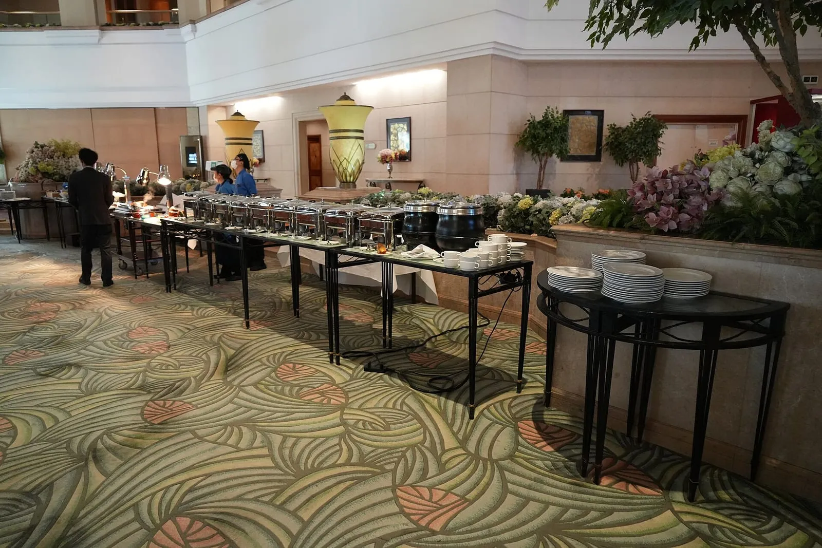Buffet dressé sur deux tables noires dans une salle de réception, plats maintenus au chaud dans des bacs en inox et piles d’assiettes blanches