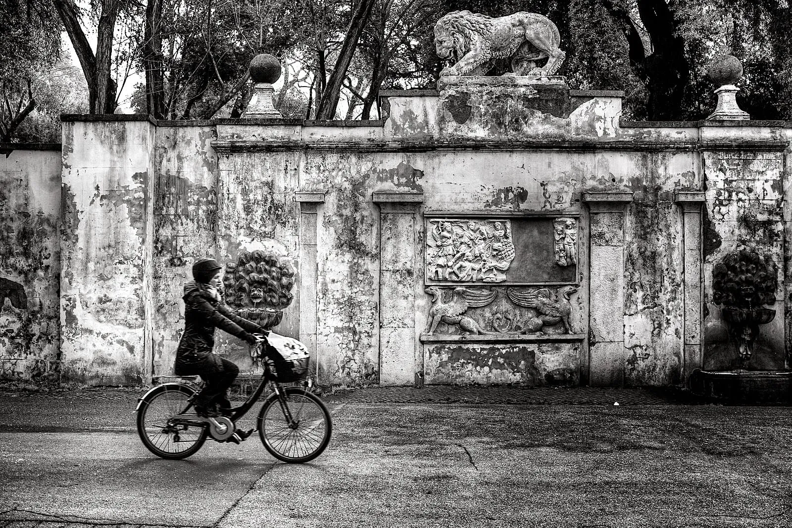 Cycliste emmitouflé passant devant un mur ancien sous un ciel d'hiver à Rome, chaussée détrempée et arbres sans feuilles