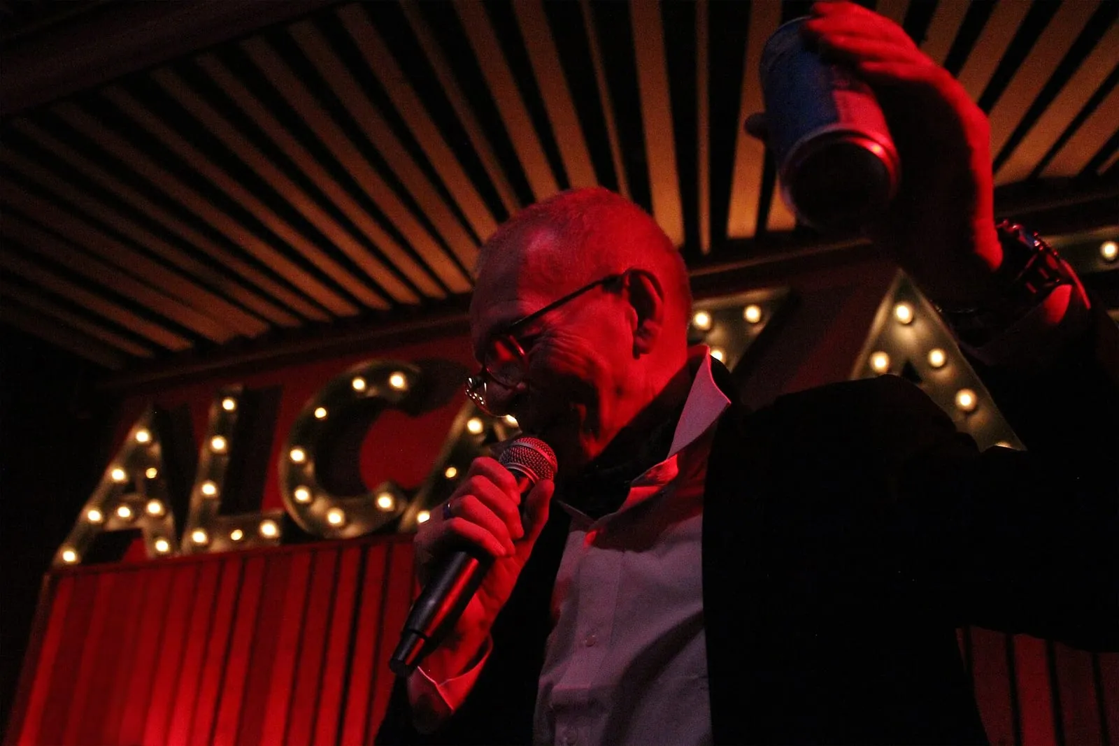 Chanteur sur la scène d'une salle de concert romaine, micro en main, sous des lettres lumineuses et un éclairage rouge