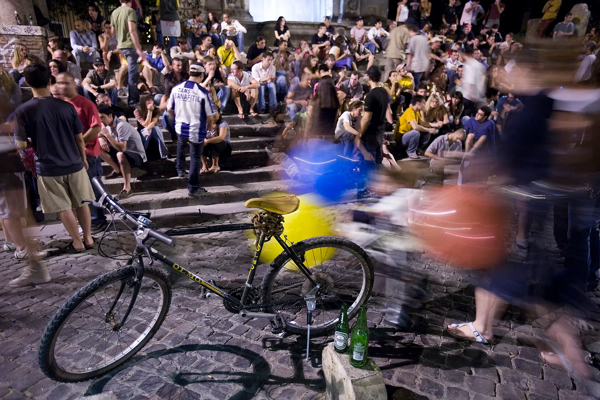 Foule assise sur les marches de la piazza Trilussa à Rome, la nuit, devant un vélo posé sur les pavés