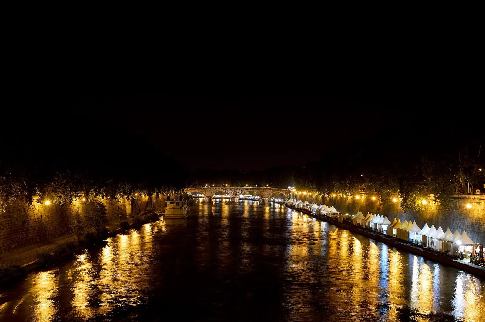 Le Ponte Sisto illuminé la nuit, vu depuis la berge du Tibre, ses arches et l'ouverture circulaire de son arche centrale reflétées dans l'eau