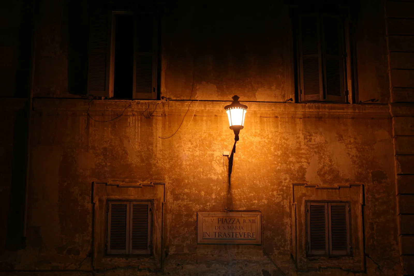 Un lampadaire allumé éclaire le mur d'un immeuble romain à la nuit tombée, au-dessus de la plaque de la piazza San Cosimato in Trastevere