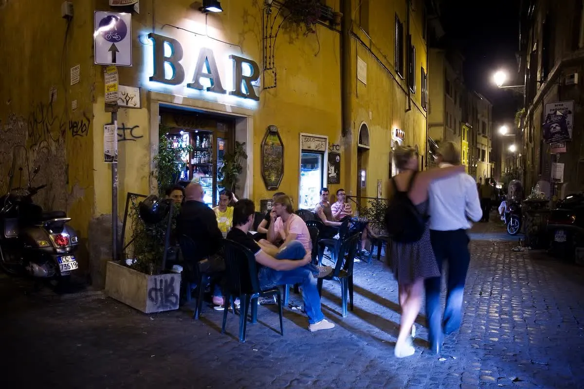 Bar d'angle du Trastevere à la nuit tombée, avec ses tables et ses chaises posées sur le pavé devant l'enseigne lumineuse