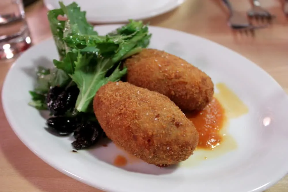 Deux supplì romains dorés et croustillants sur une assiette, avec olives noires et salade